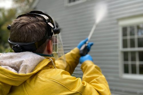 pexels-photo-5652626-5652626 Person cleaning house siding with a pressure washer, wearing protective gear in a yellow raincoat.
